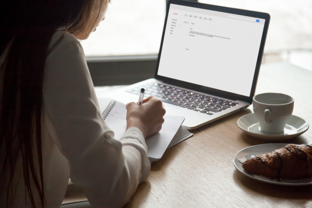 Mulher sentada em um café olhando uma planilha em um laptop. Ao lado do computador, há uma xicará de café em um pires, e um prato com um croissant.